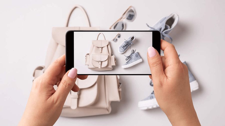 Hands holding a smartphone to photograph a beige backpack, sunglasses, and blue sneakers laid out.