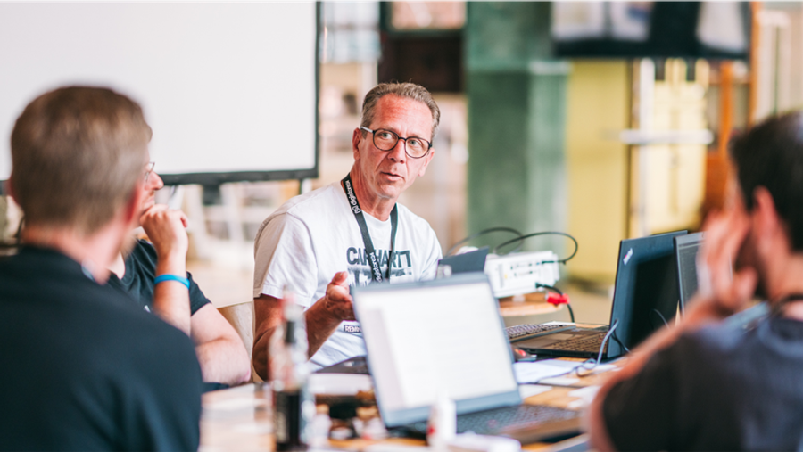 A man in a white t-shirt and glasses speaks at a table with laptops during a meeting.