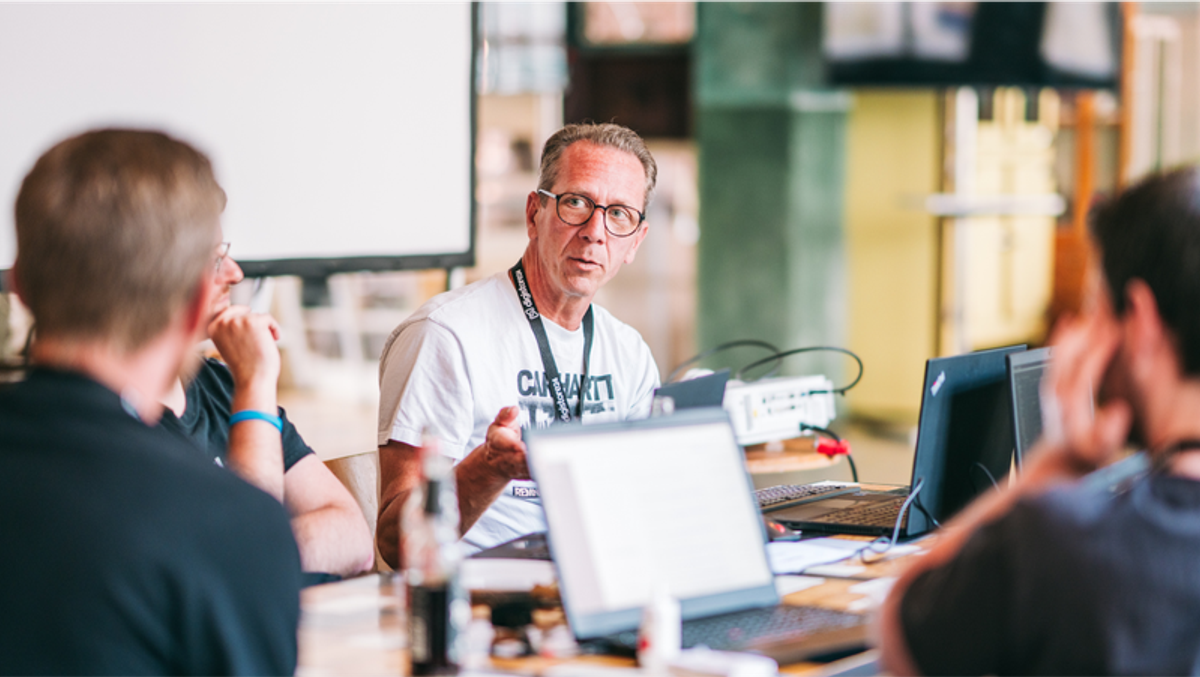 A man in a white t-shirt and glasses speaks at a table with laptops during a meeting.
