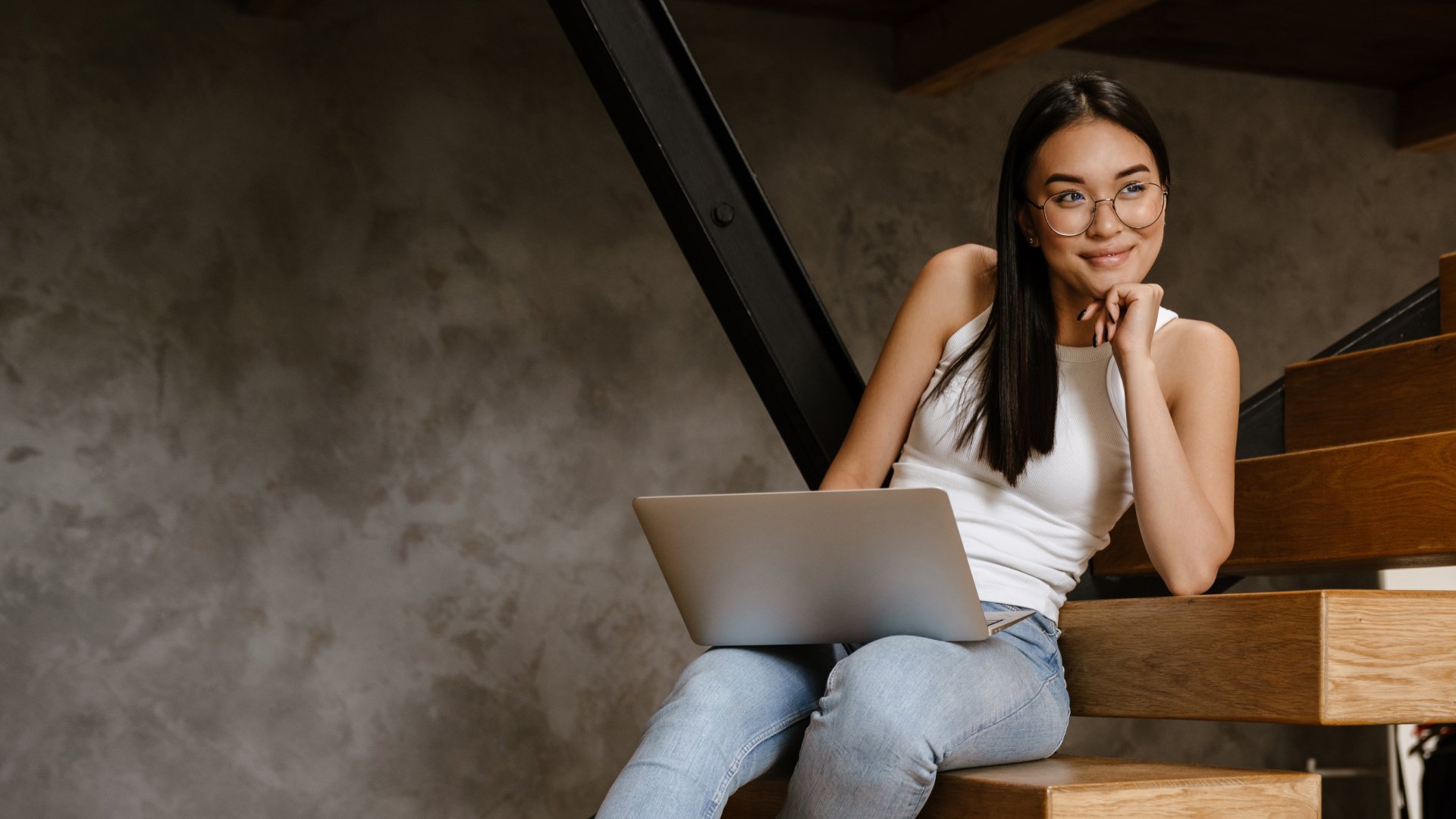 Jovem mulher asiática sentada em escadas de madeira com um laptop, sorrindo e olhando para o lado.