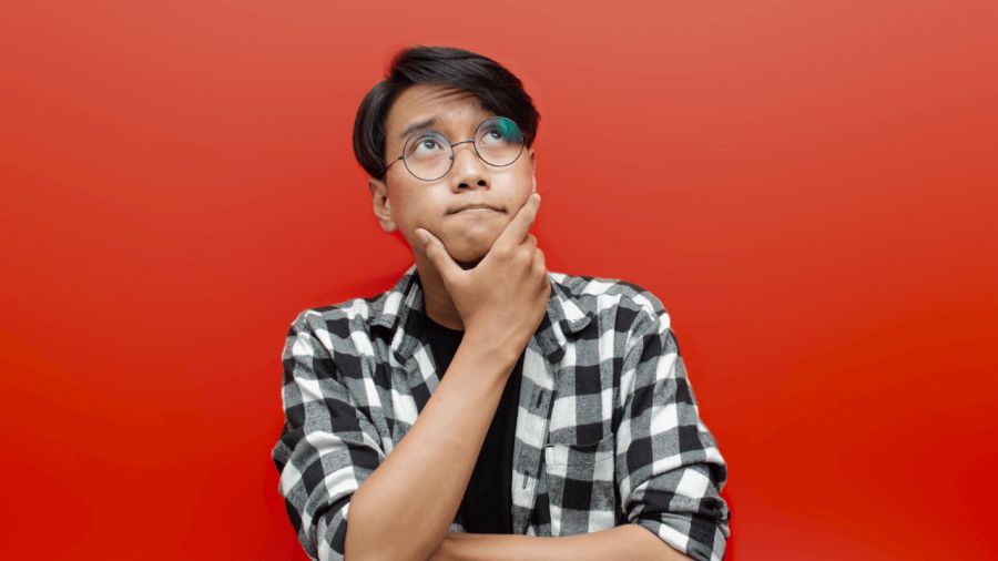 Thoughtful young man in glasses and plaid shirt looks up, hand on chin, against a red background.