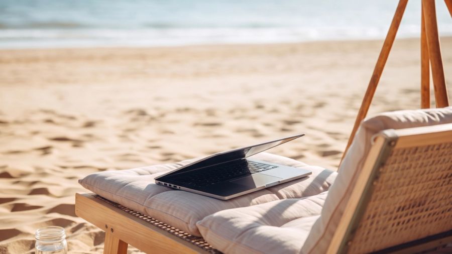 Person typing on a laptop at a table overlooking a sunny beach and ocean.