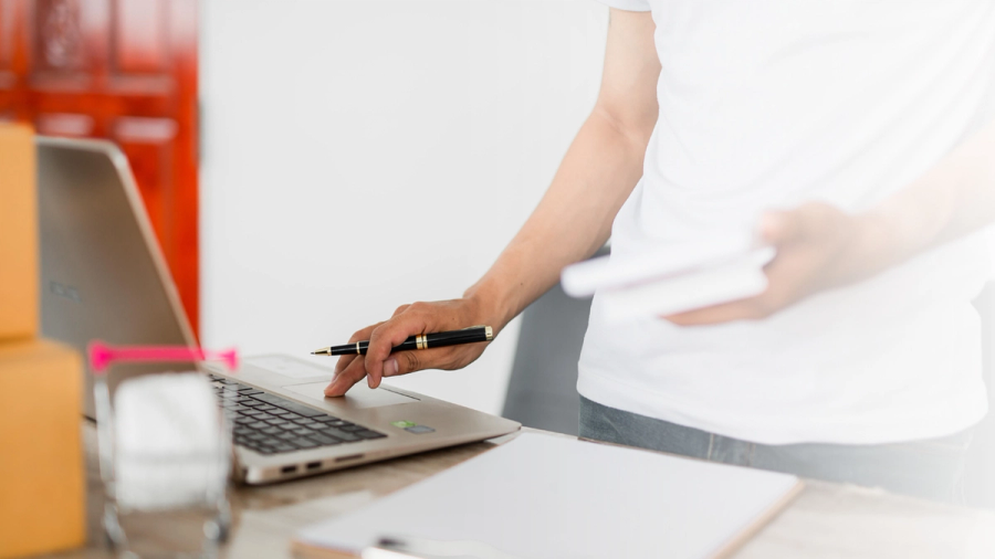 A person works on a laptop at a desk, holding a pen and papers, with shipping boxes nearby.
