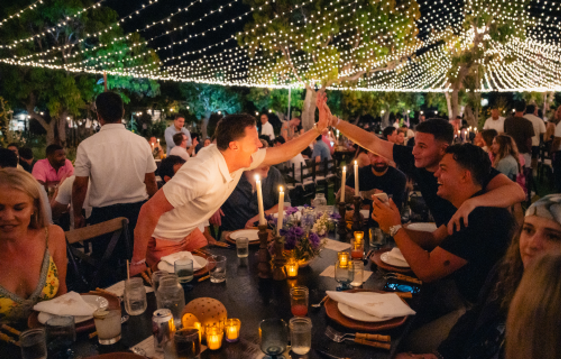 Two men high-fiving at a lively outdoor dinner party at night, lit by string lights and candles.