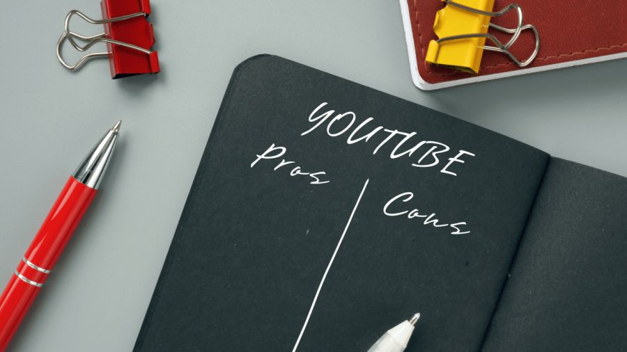 Black notebook titled "YOUTUBE Pros Cons" with a red pen and binder clips on a grey surface.