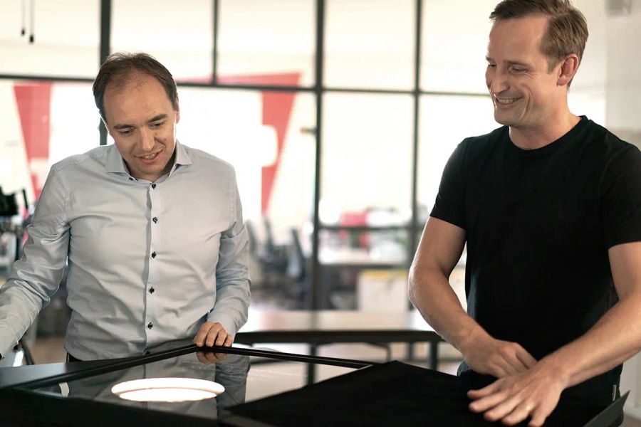 Two men engaging with a glowing circular display on a black table, one smiling.