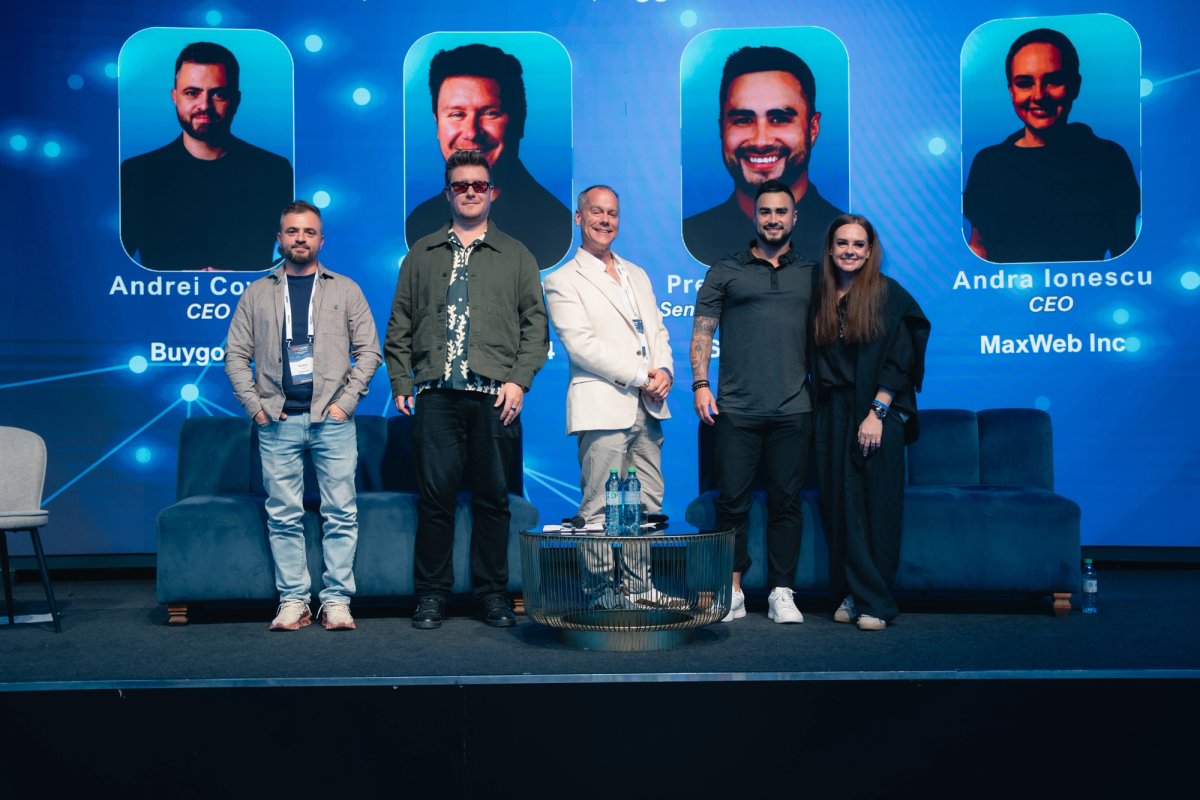 Five panelists, four men and one woman, stand on a stage in front of a blue screen displaying their headshots and professional titles.