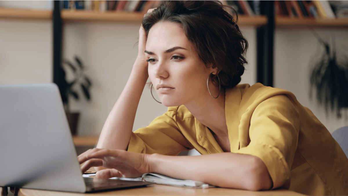 A woman with a tired expression looks at a laptop, resting her head on her hand.