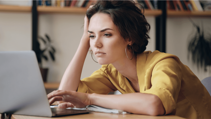 A woman with a tired expression looks at a laptop, resting her head on her hand.