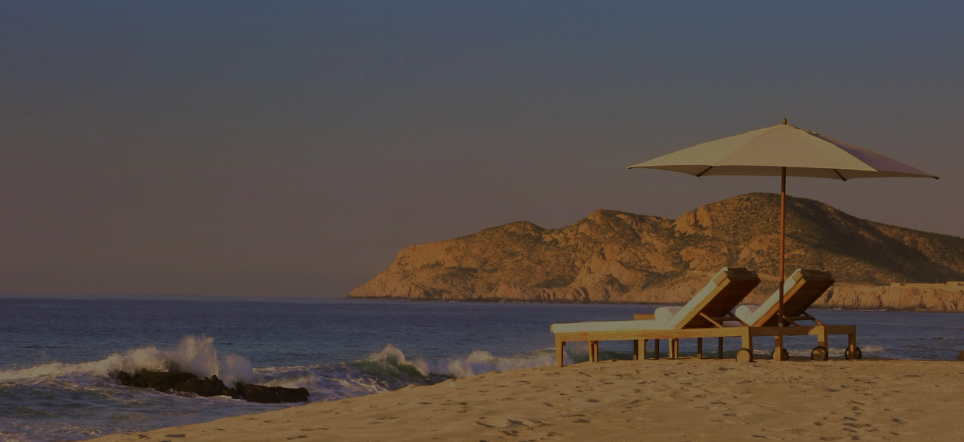 Two beach chairs and an umbrella on a sandy shore with ocean waves and mountains.