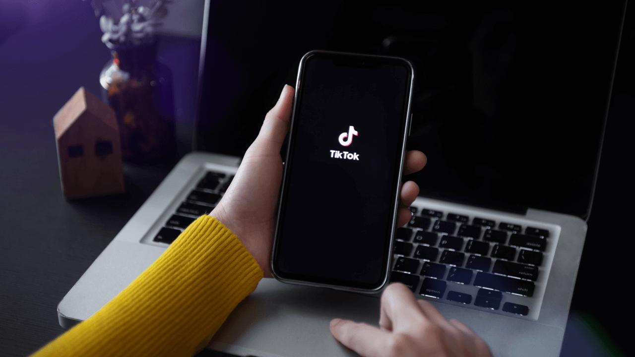 A hand holds a smartphone displaying the TikTok logo, with a laptop keyboard in the background.