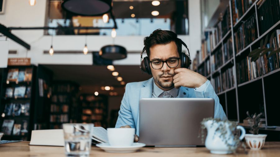 A man wearing headphones and glasses intently uses a laptop in a cafe or library.