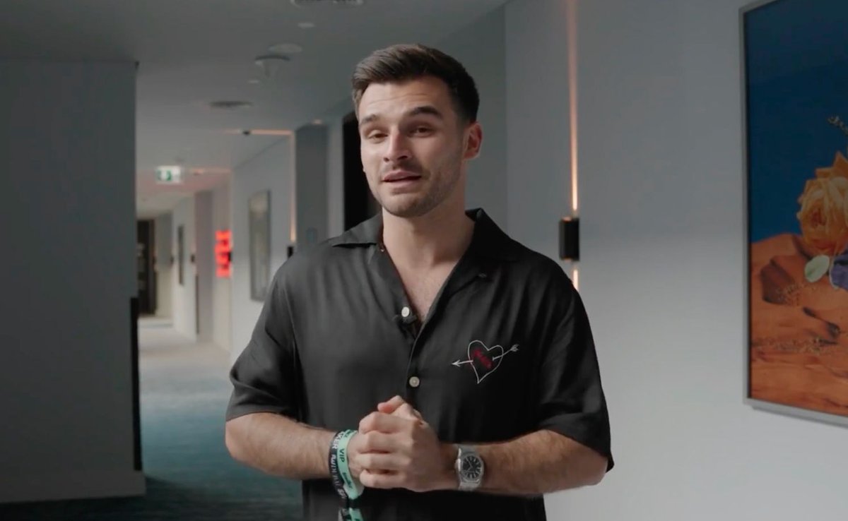 A man in a black shirt with a heart-and-arrow design stands in a hallway.