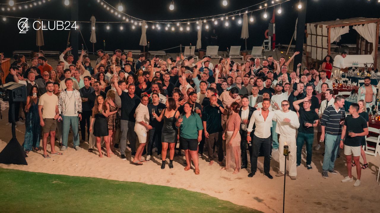 A large group of people smile and pose on a sandy beach at night under string lights, with the CLUB24 logo.