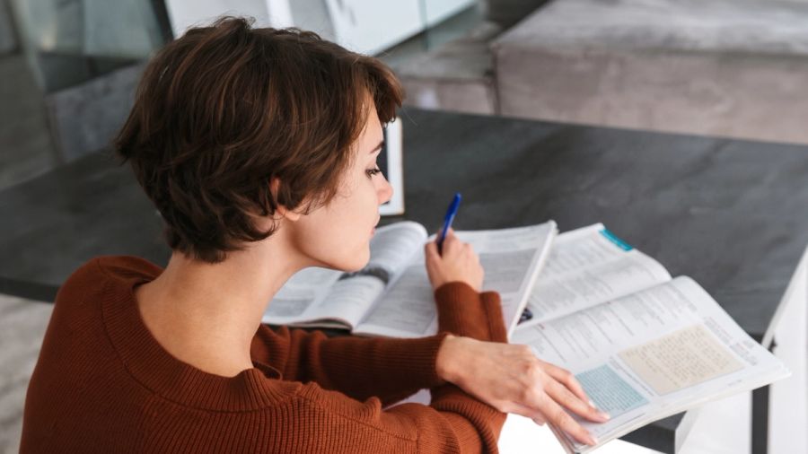A woman with short brown hair studies from an open textbook, holding a blue pen.