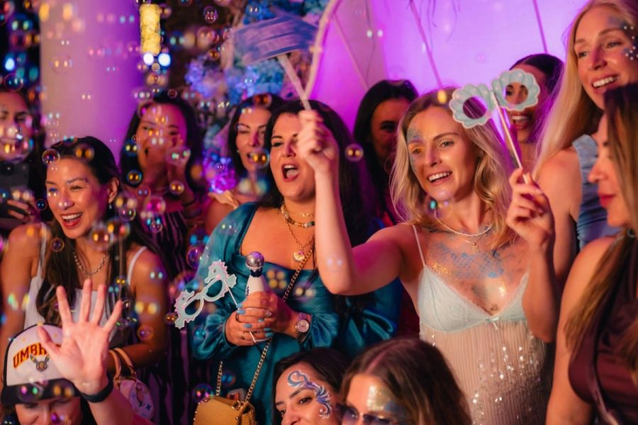 A diverse group of women smiling and laughing at a vibrant party with bubbles, some holding props.