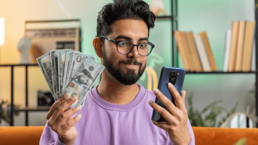 A man with a surprised expression holds a stack of cash and a smartphone.