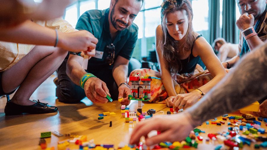 A group of people collaboratively building with colorful LEGO bricks on a wooden floor.