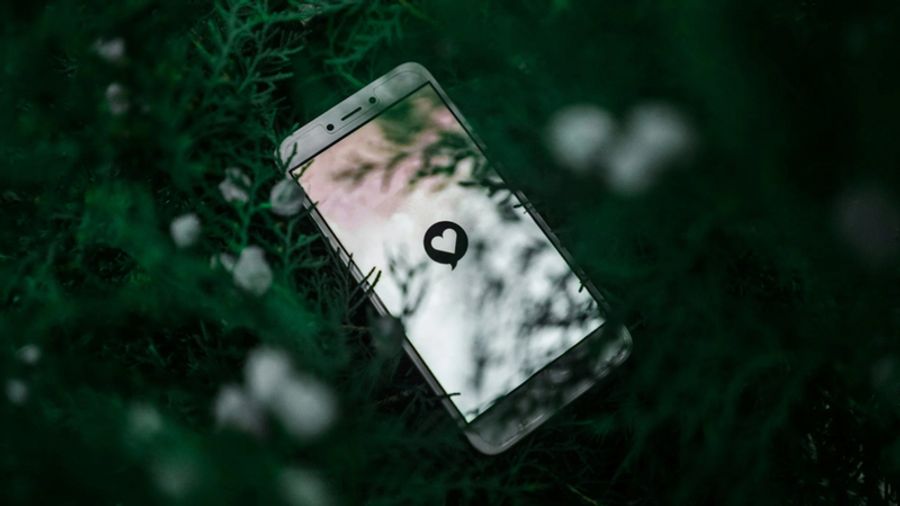 A white smartphone rests on dark green foliage, its screen showing a black heart in a speech bubble.