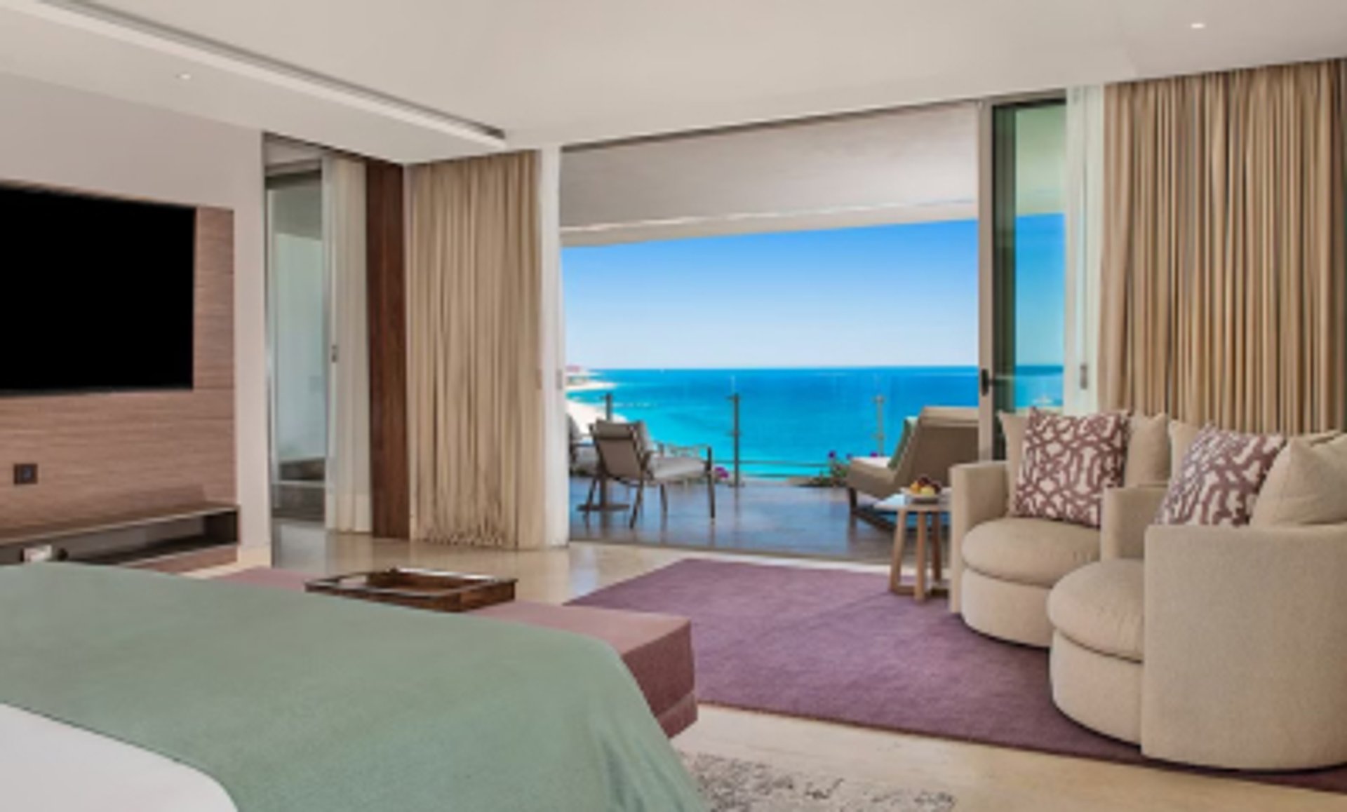 A luxurious bedroom with a large bed, TV, and armchairs, opening onto a balcony with stunning views of a turquoise ocean.