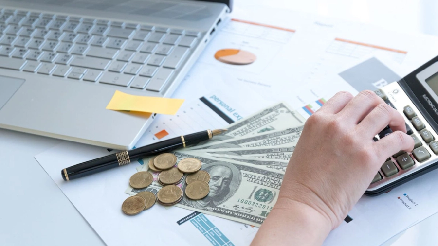 A hand uses a calculator amidst financial documents, cash, coins, and a laptop.