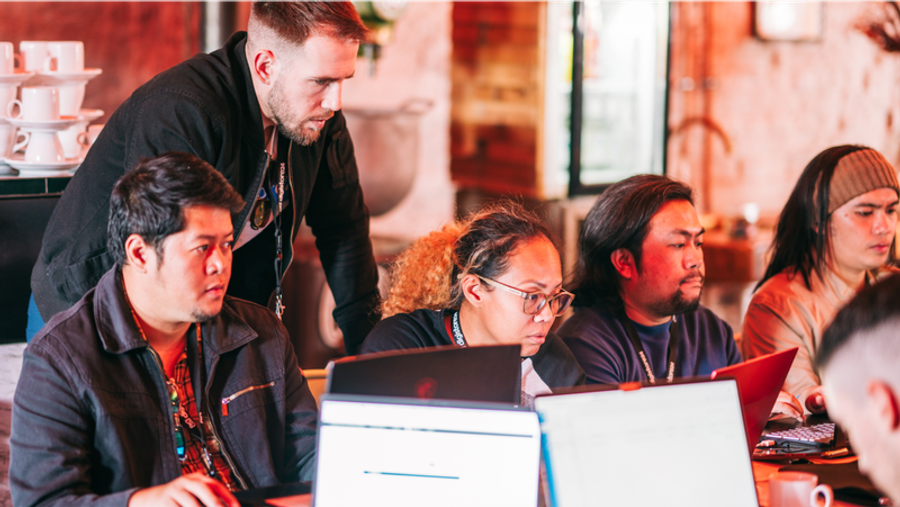 A diverse group of people intensely focused on laptops, with one man standing and observing another's screen.