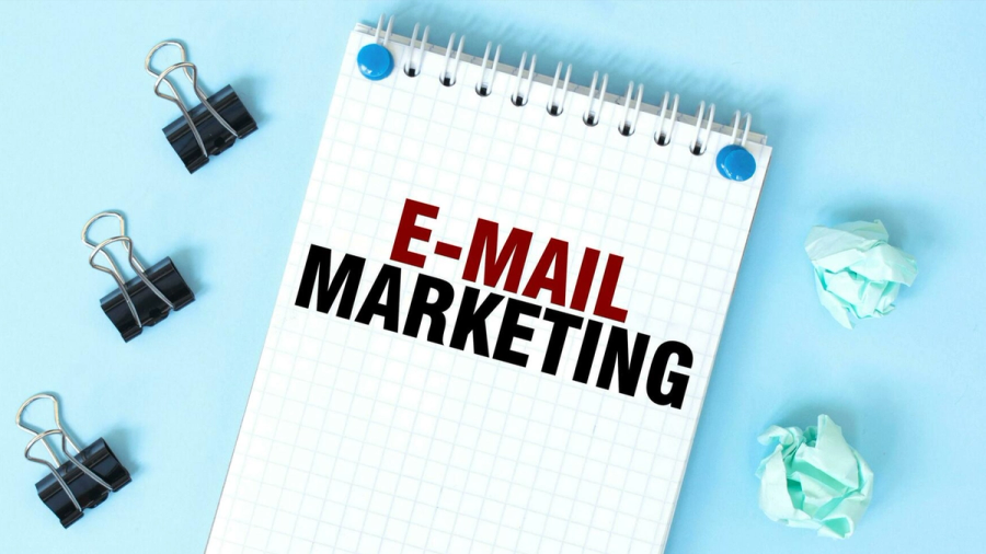 A notepad with "E-MAIL MARKETING" written on it, surrounded by binder clips and crumpled paper on a blue surface.
