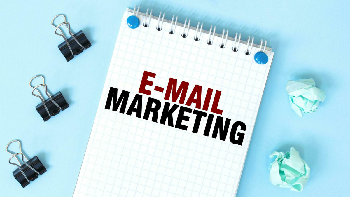 A notepad with "E-MAIL MARKETING" written on it, surrounded by binder clips and crumpled paper on a blue surface.