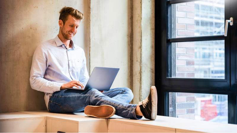 A smiling young man sits cross-legged on a bench while working on a laptop.