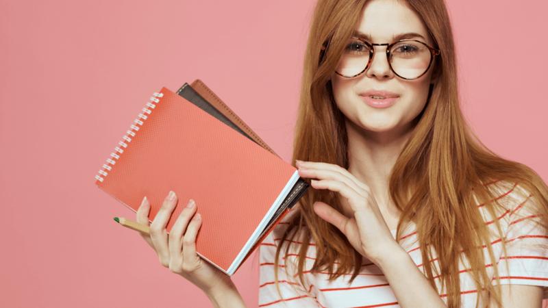A woman with glasses and long hair smiles, holding spiral notebooks and a pencil.