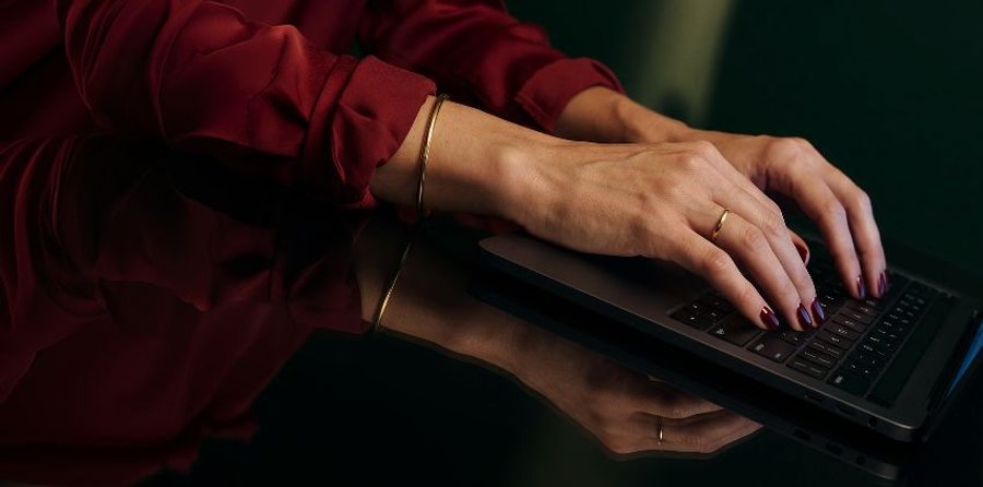 Hands in a red shirt with dark nail polish typing on a laptop.