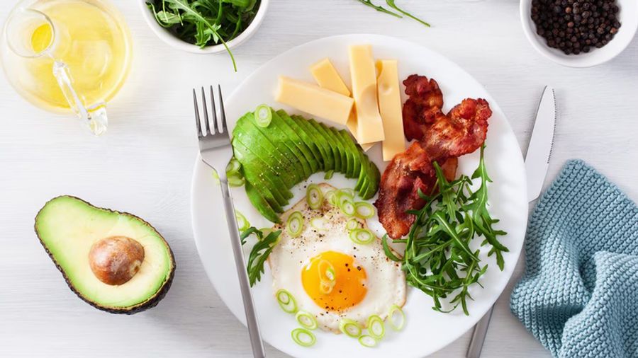A white plate with a fried egg, fanned avocado slices, crispy bacon, cheese, and arugula.