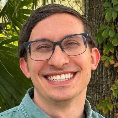 A smiling man wearing glasses against a leafy outdoor background.