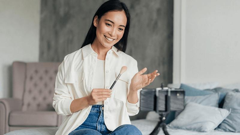 A smiling Asian woman holding a makeup brush, recording a video with a smartphone on a tripod.