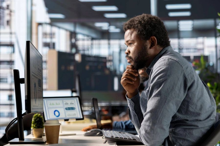 A Black man intently views computer screens displaying data in an office.