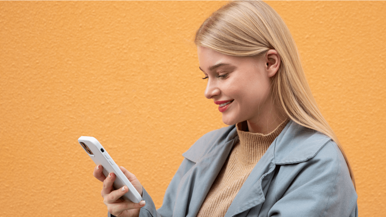 Young blonde woman smiling while looking at a smartphone against an orange wall.