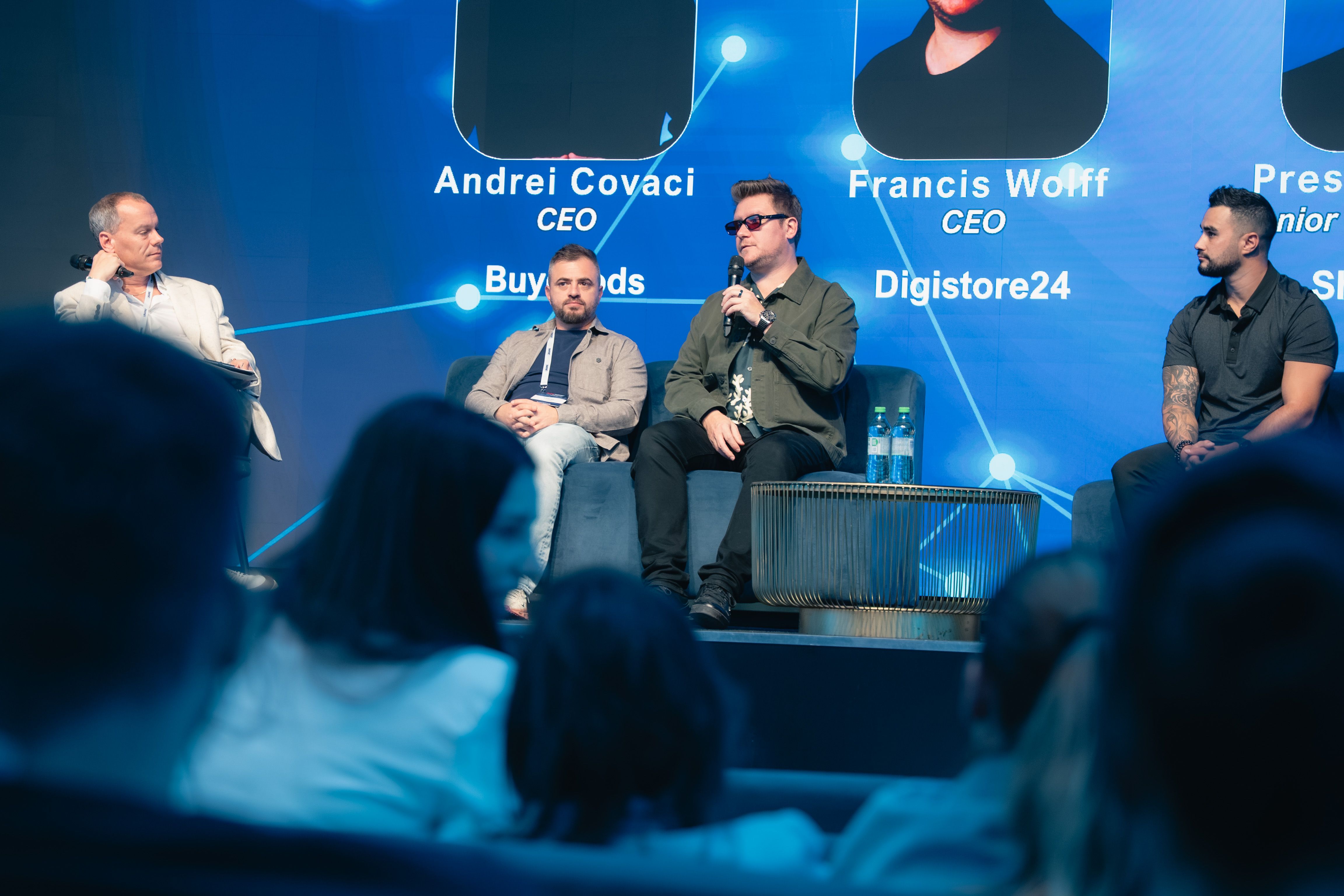 Four men on stage for a panel discussion; one speaks into a microphone. A blue screen behind them displays names like Francis Wolff, CEO Digistore24. An audience sits in the foreground.