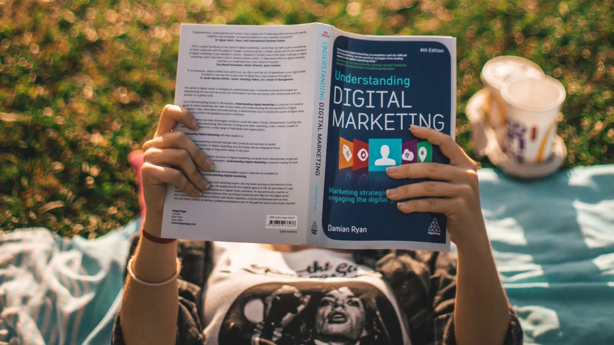 Person lying on a blanket on grass reading a book titled "Understanding Digital Marketing".