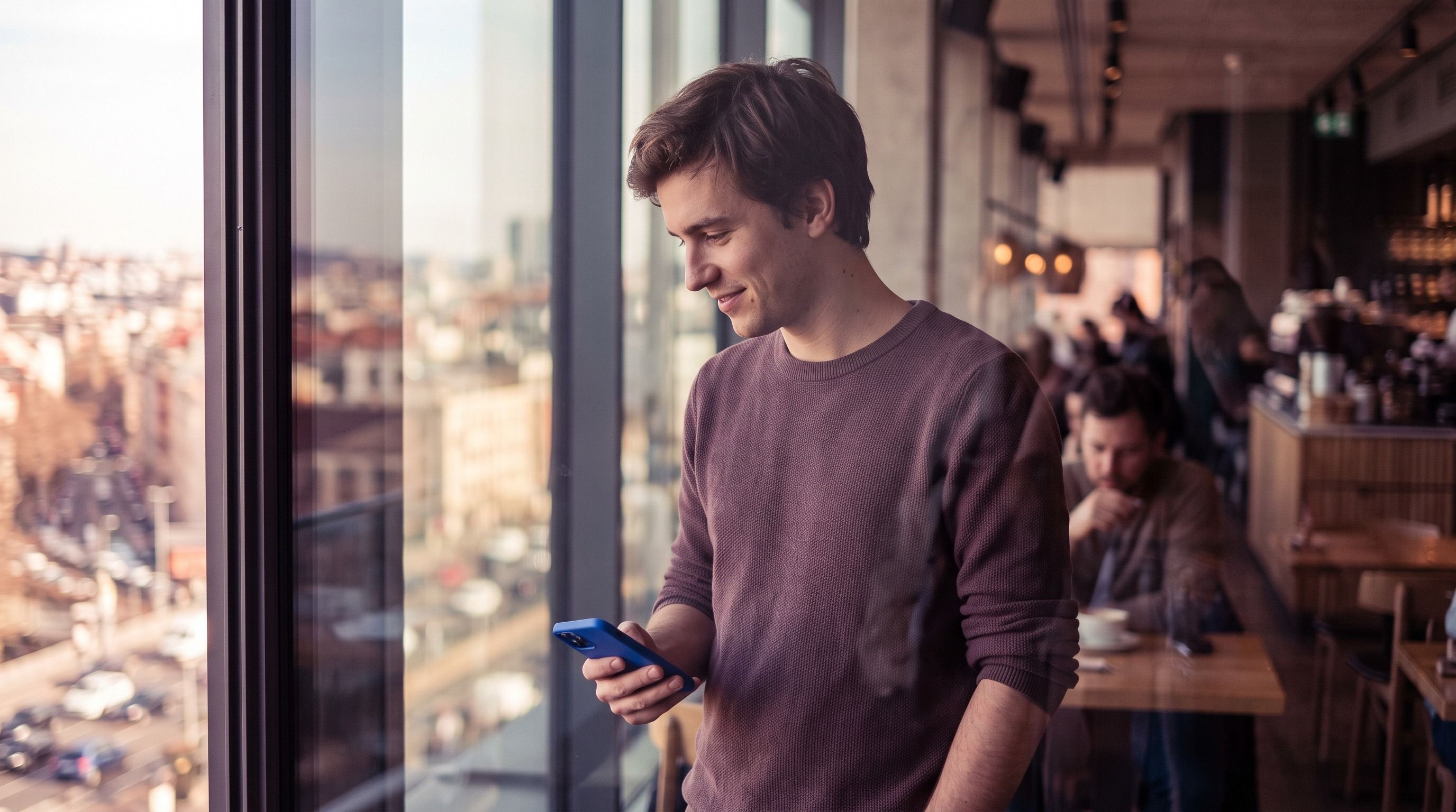 Young man smiles while looking at his phone by a window with a city view and cafe background.