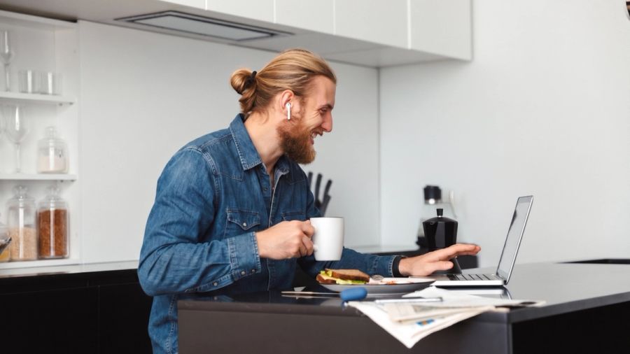 Smiling man with earbuds using a laptop at a kitchen counter, holding a mug, with a sandwich nearby.