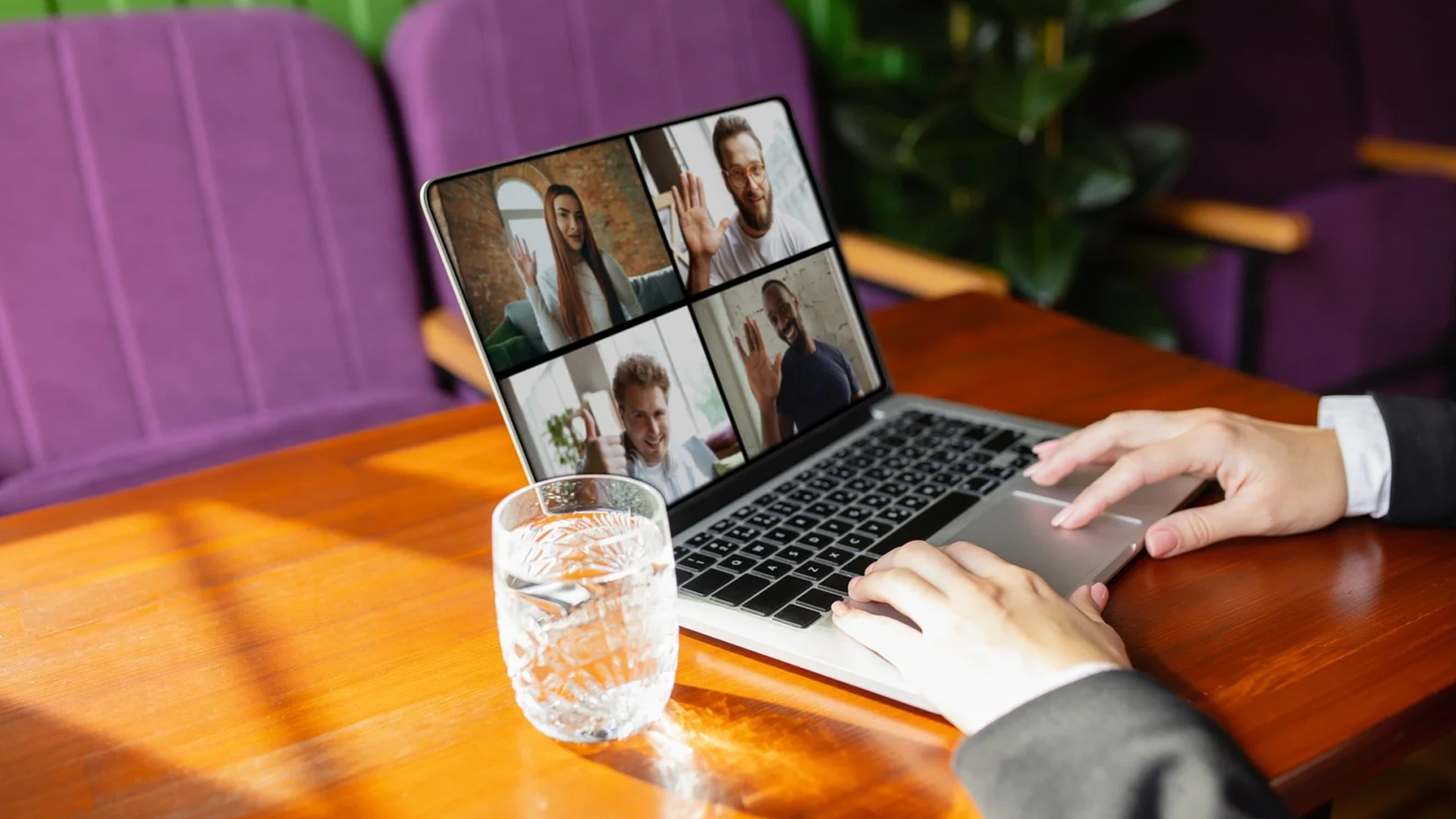 Hands using a laptop displaying a four-person video conference, with a glass of water on a wooden table.