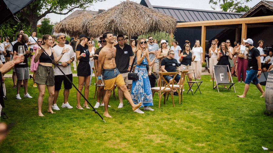 A crowd of people gathered on a green lawn, applauding as a shirtless man in shorts smiles brightly, with a building and thatched roof in the background.