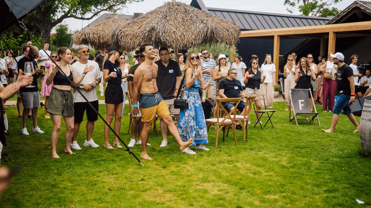 A crowd of people gathered on a green lawn, applauding as a shirtless man in shorts smiles brightly, with a building and thatched roof in the background.