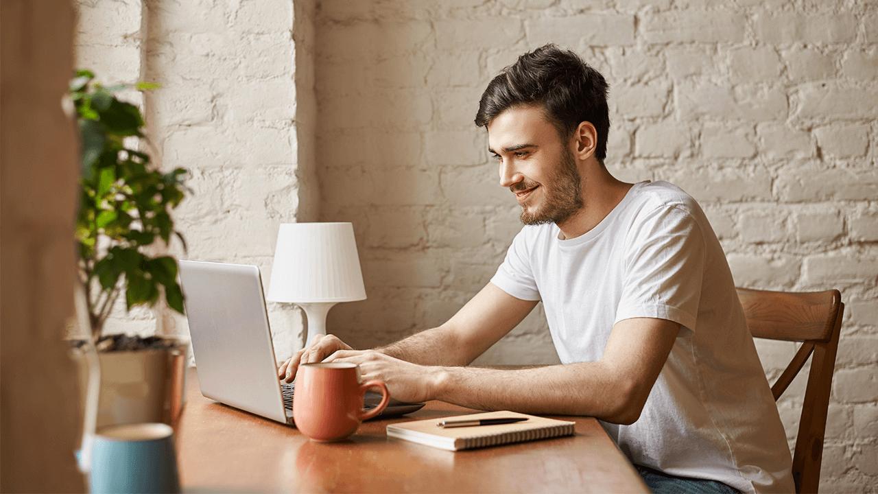 A smiling man in a white t-shirt types on a laptop at a wooden table with a mug and notebook.