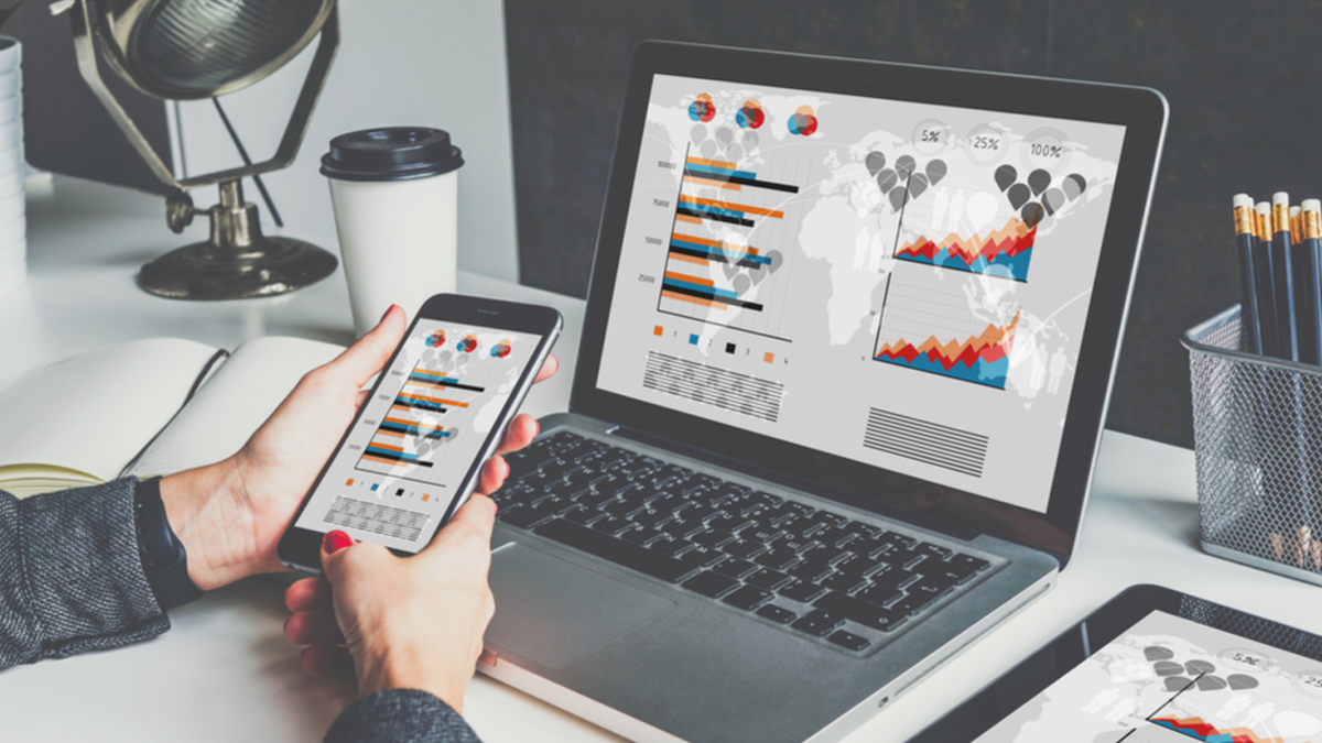 A person's hands hold a smartphone displaying data charts, next to a laptop and tablet with similar data visualizations.