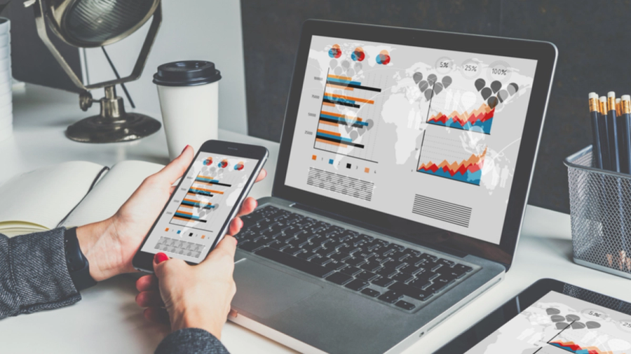 A person's hands hold a smartphone displaying data charts, next to a laptop and tablet with similar data visualizations.