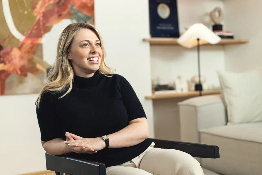 A smiling blonde woman in a black top sits in a chair with hands clasped.