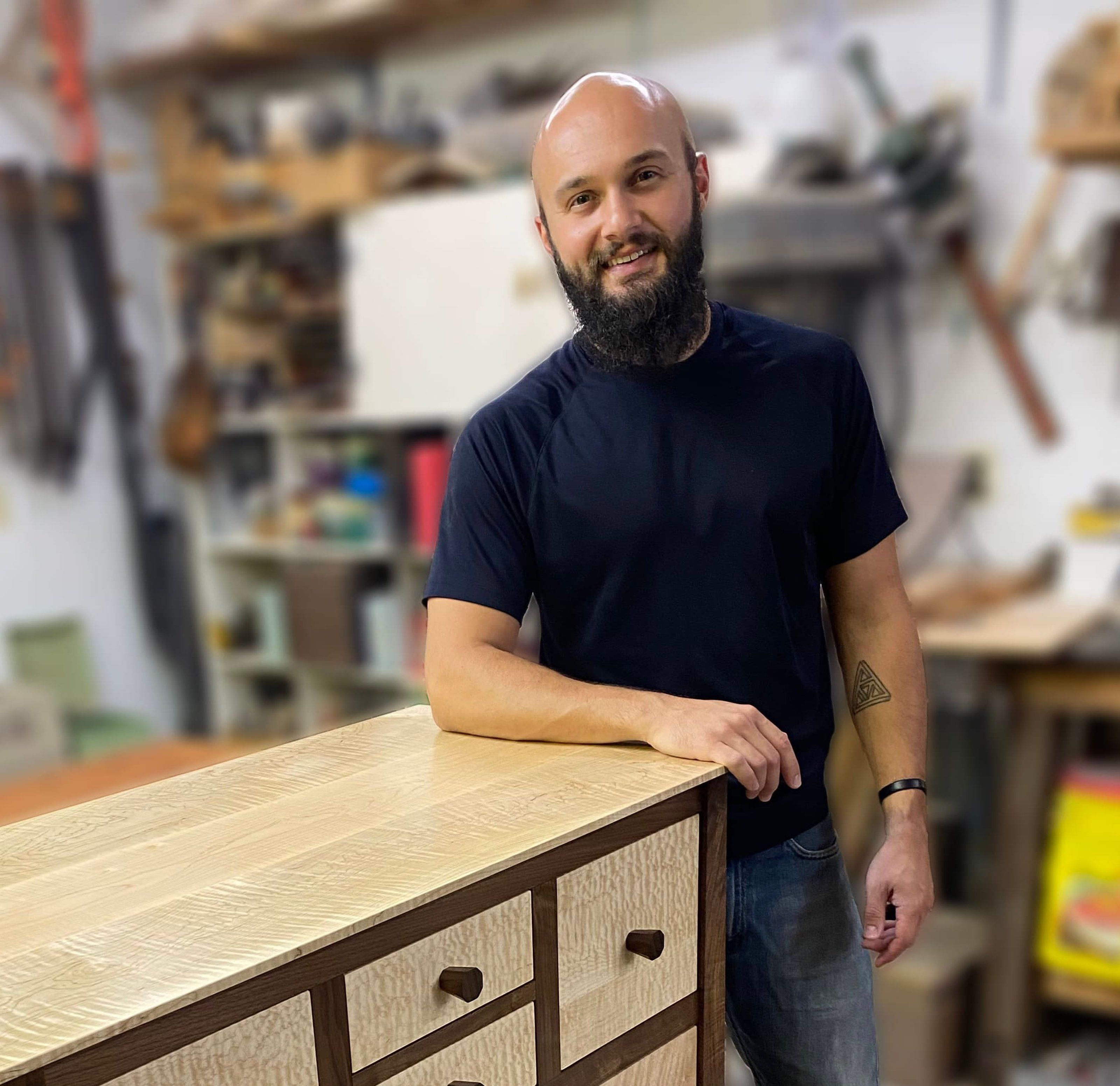 A smiling, bald, bearded man leans on a two-toned wooden cabinet with light, textured drawers and a dark frame, in a blurred workshop.