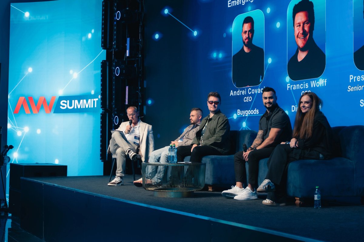 Five panelists sit on blue couches on stage at the AW Summit, with one man speaking into a microphone.