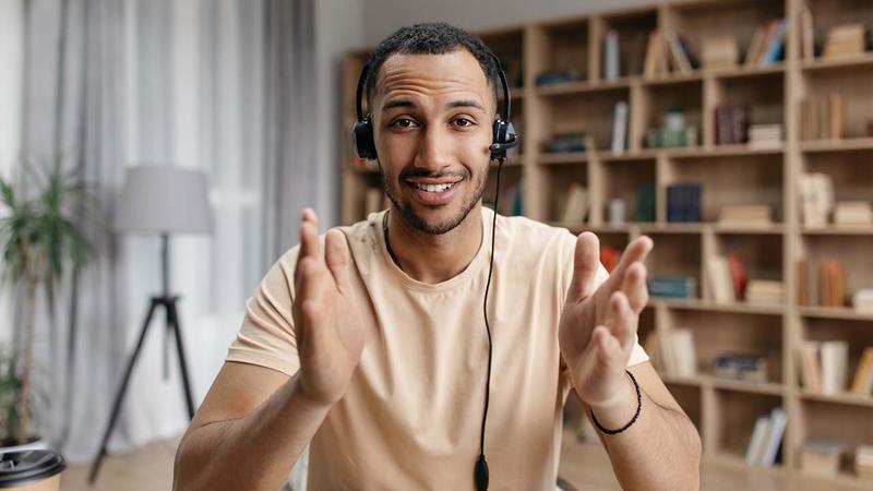 A smiling man wearing a headset gestures with open hands while looking at the camera.